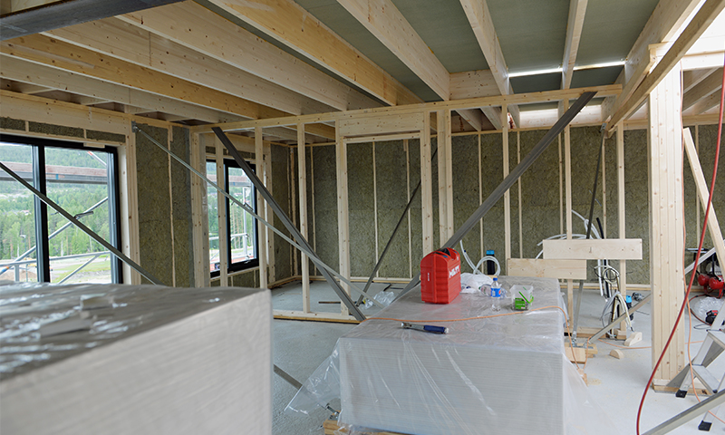 Byggeplass inne med lyse trekonstruksjoner, grønn isolasjon, metallstag og rød verktøykasse.