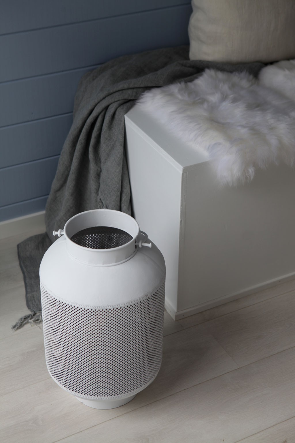 Perforert hvit metalllykt på lyst tregulv foran hvit kasse med pels og grått pledd mot blå vegg