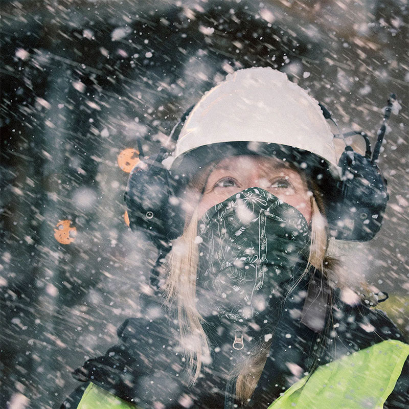 Person med hvit hjelm, svarte hørselsvern og refleksvest i tett snøvær i skog.