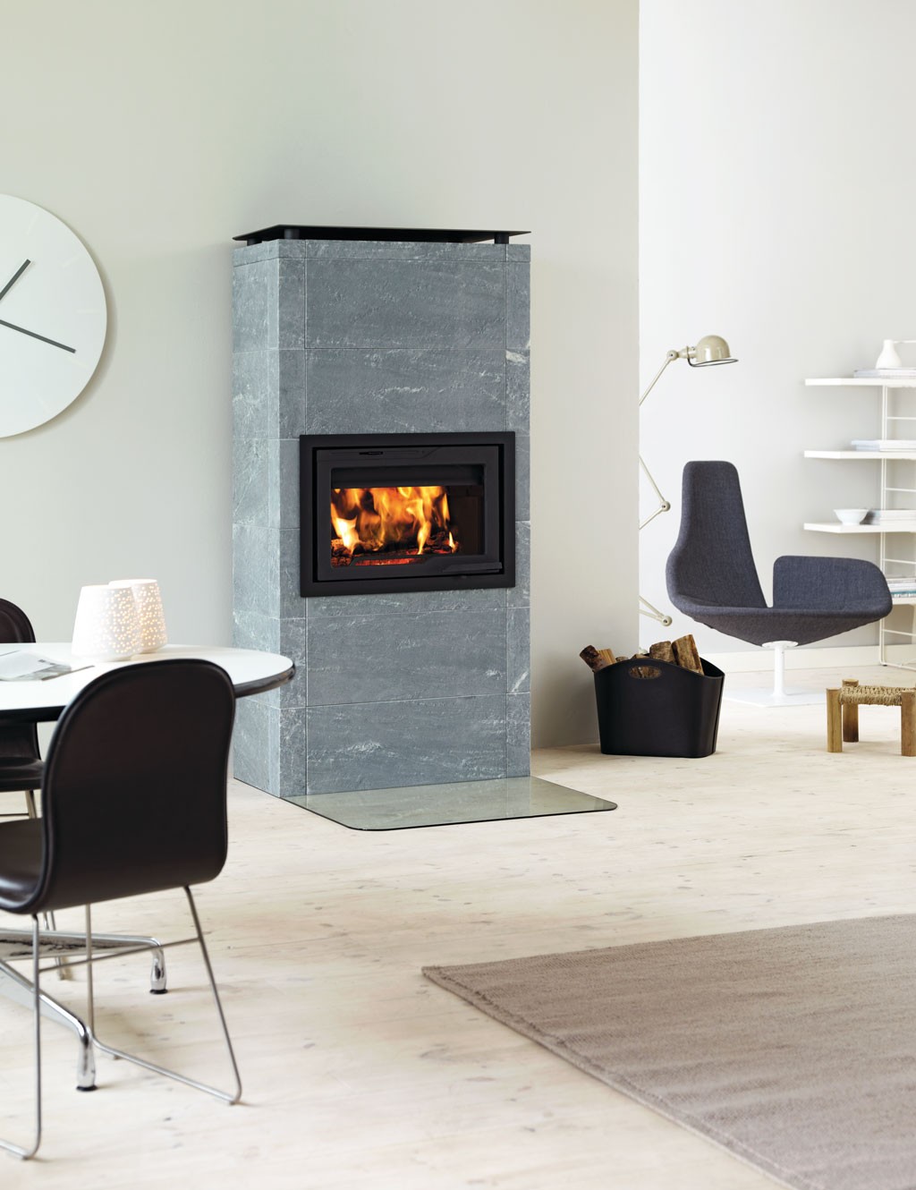 Moderne lyst rom med grå klebersteinspeis med flamme, vedkurv, hvit veggur og grå lenestol