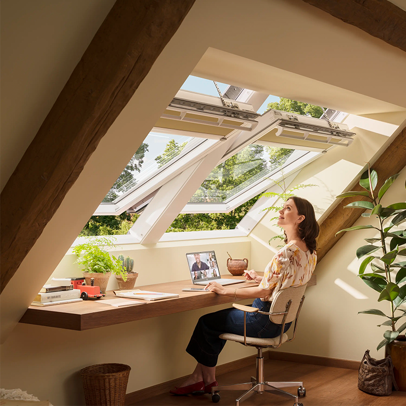 Kvinne jobber ved skrivebord på loft med åpne takvinduer, naturlig lys og grønne planter rundt seg.