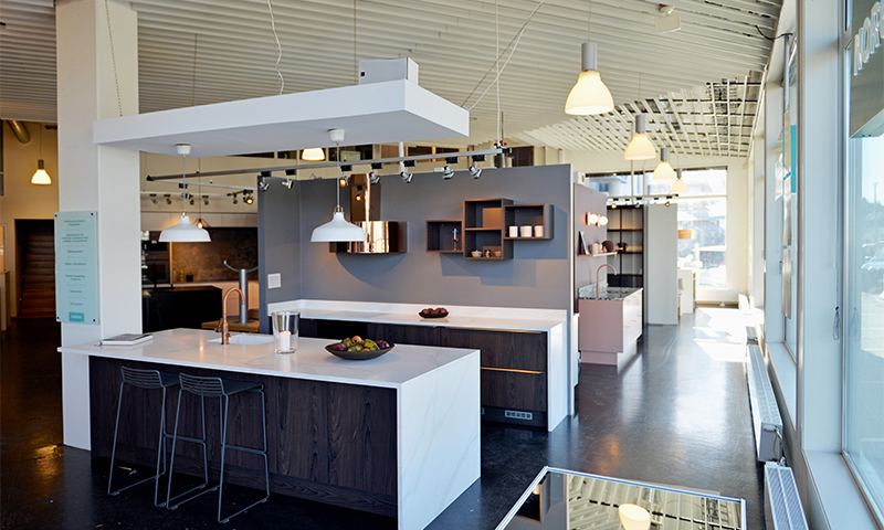 Moderne kjøkkenutstilling med hvite benkeplater, mørke treskap, grå vegg og pendellamper.