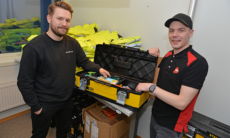 To menn ved åpen gul-svart verktøykasse i rom med stabler av gule vernevester.