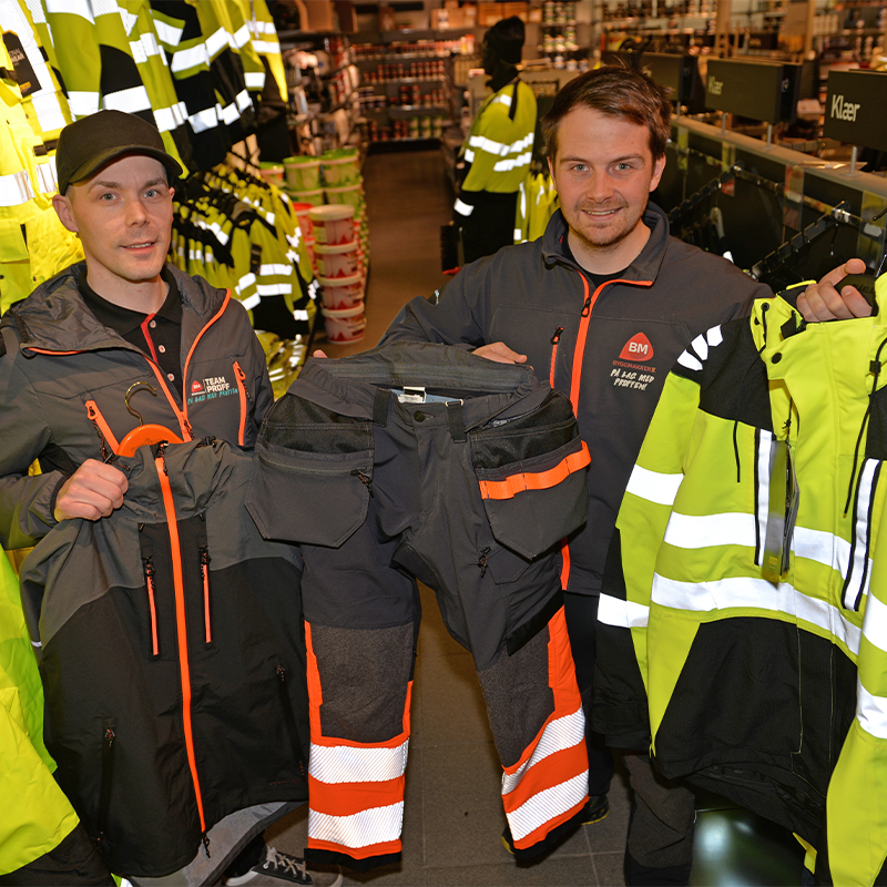 To ansatte i butikk holder gul-svart refleksjakke og grå bukse med oransje detaljer.