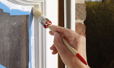 Hånd maler hvit vinduskarm med pensel, blå malertape på kanten og murvegg bak.