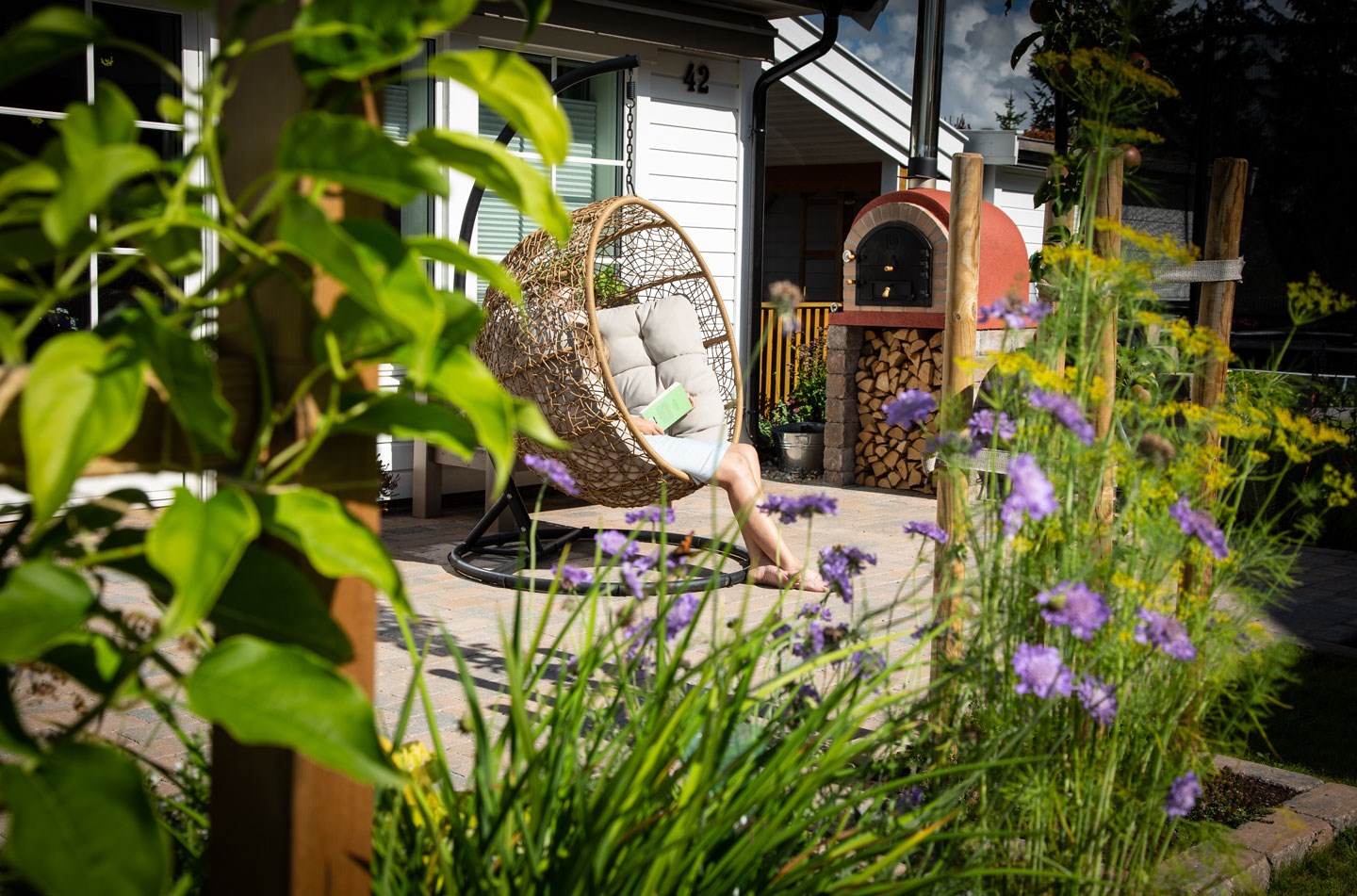 Person leser i kurvformet hengestol på lys terrasse med lilla blomster og vedovn.