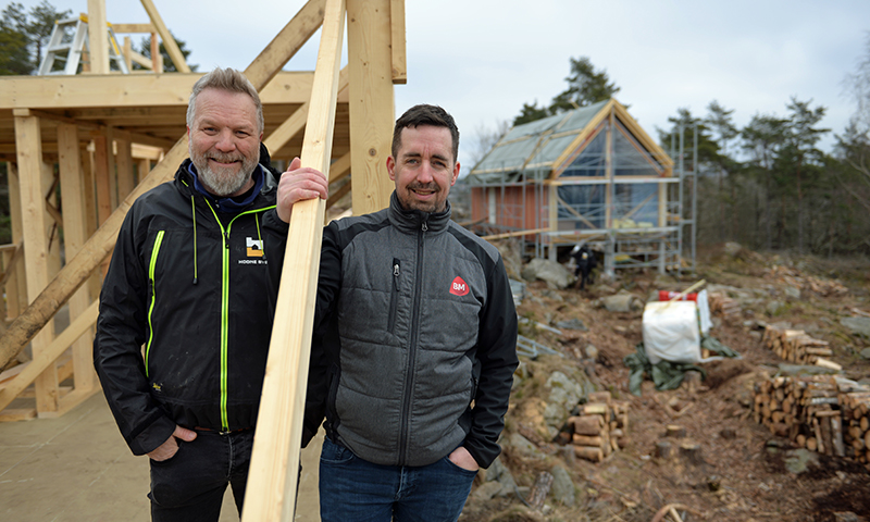 To håndverkere i svarte og grå jakker holder en planke foran nytt trehus under bygging.