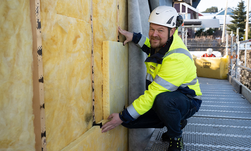 Arbeider i gul refleksjakke og hvit hjelm installerer gul isolasjon på husvegg fra stillas