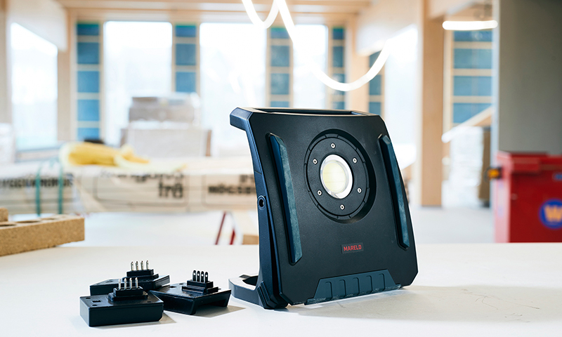 Svart bærbar arbeidslampe med rund LED på byggeplass, tre ekstra batteripakker på bordet