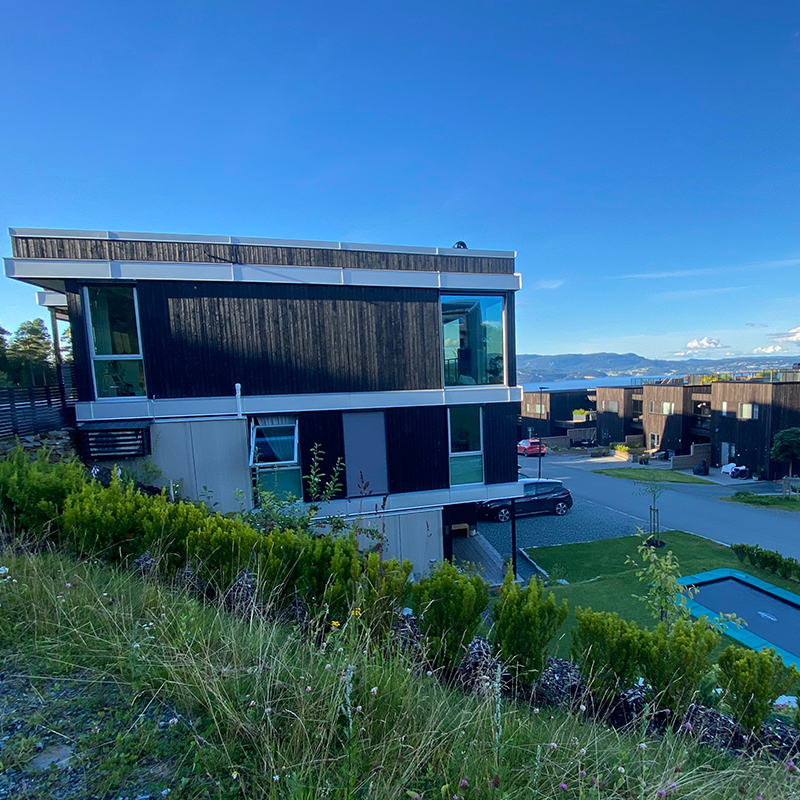 Moderne boligblokk i mørkt treverk og hvitt, foran grønn skråning, biler og blå fjordutsikt.