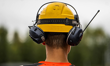 Mann bakfra med gul hjelm, svart hørselsvern og oransje vest foran uskarp skog