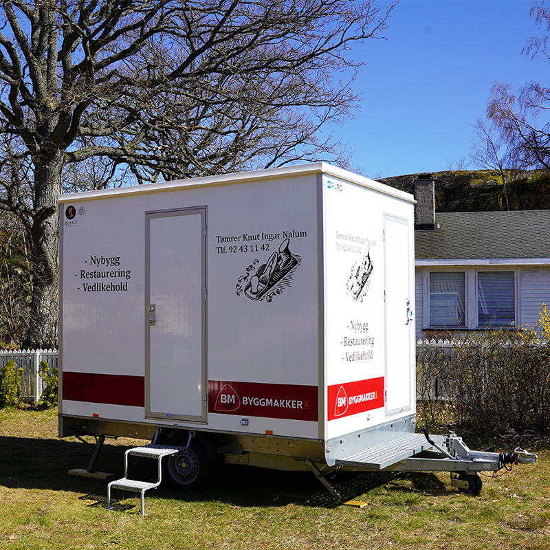 Hvit mobil byggebod med rød Byggmakker-logo og tømrerreklame, plassert på gress i hage.