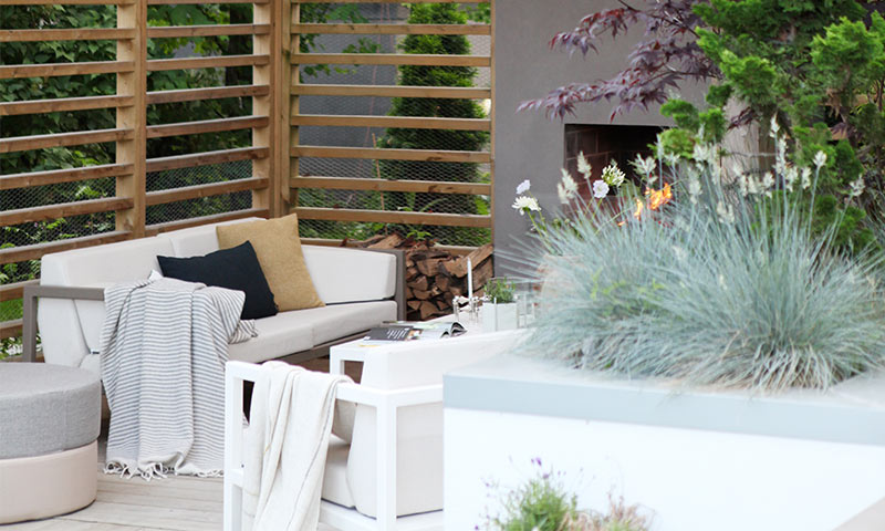 Uteplass med hvit sofa, sorte og okergule puter, pledd, gressplanter og tent peis.