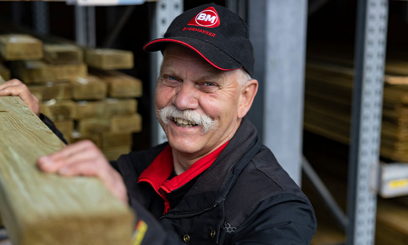 Ansatt i svart og rødt BM Byggmakker-tøy løfter treplanke foran lagerhylle.