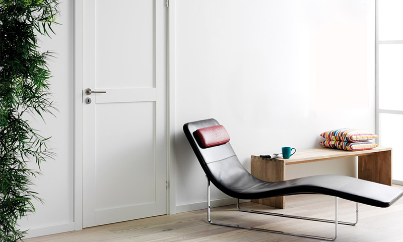 Svart lenestol i stue med rød nakkestøtte ved hvit dør, trebenk med fargerike puter og grønn kopp.