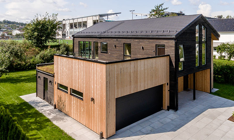 Moderne toetasjes hus i mørkt og lyst trepanel med store vinduer, dobbelgarasje, grønn plen