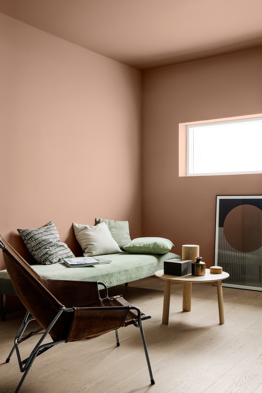 Minimalistisk stue med ferskenfargede vegger, grønn daybed, brun lærstol og rundt trebord.