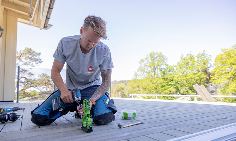 Håndverker på kne monterer grå terrassebord med drill og spikerpistol i hage.