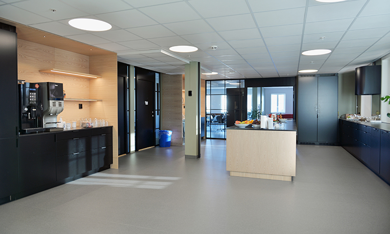 Lyst pauserom med sorte skap, lys kjøkkenøy, kaffemaskin, fruktfat og glassvegger.