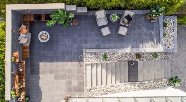 Dronebilde av grå flislagt terrasse med lounge-stoler, planter og trapp omgitt av grønthekk.