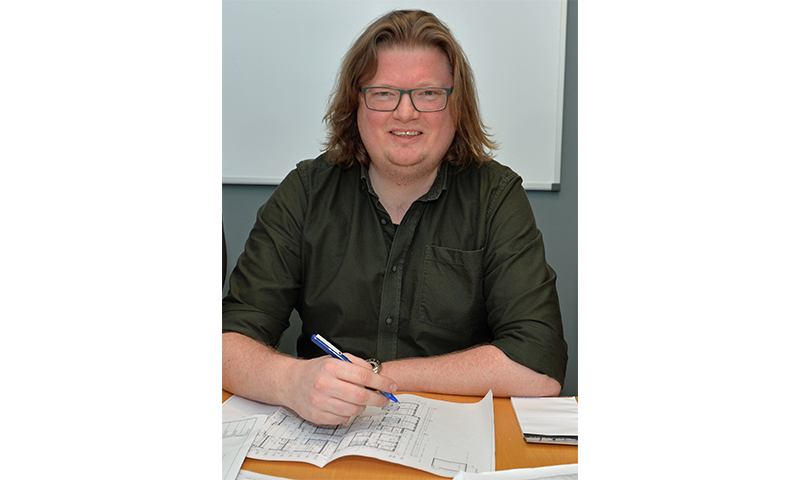 Mann med grønn skjorte og skulderlangt hår tegner på plantegninger på bord foran hvit tavle.