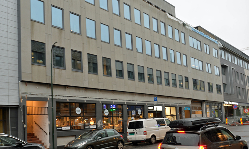 Beige fleretasjes kontorbygg med butikkvinduer, parkert hvit varebil og småbiler under grå himmel