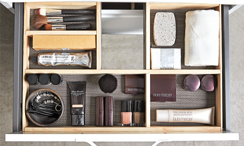 Åpen skuff med treinndelte rom, sminkekoster, kremer, svamper, neglelakk på grå bakgrunn.