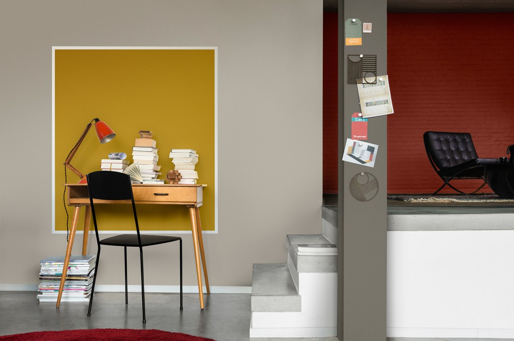 Hjemmekontor med trebord dekket av bøker foran gul firkant på grå vegg, rød bordlampe og svart stol