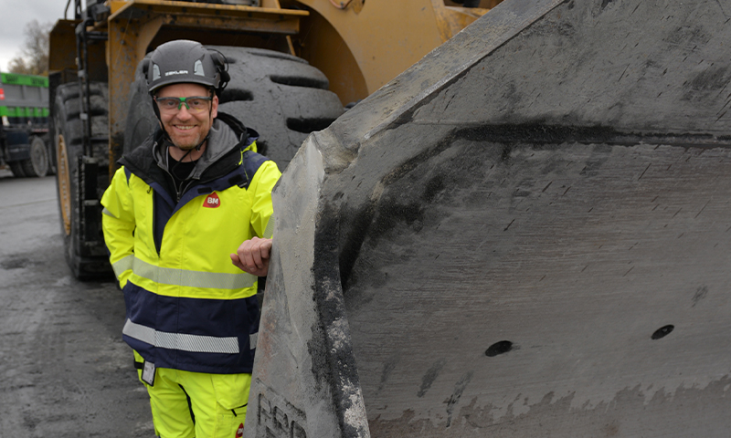 Arbeider i gul refleksjakke og hjelm ved stor gul hjullasterskuffe på asfaltplass