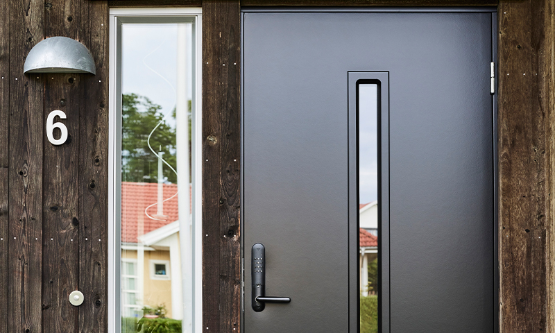 Moderne svart inngangsdør i mørkt trepanel med smalt vindu, utsikt til gult hus med rødt tak.