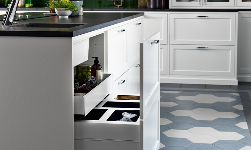 Hvit kjøkkenøy med svart benkeplate, åpnede skuffer for avfallssortering, heksagonfliser på gulvet.