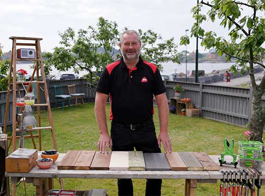 Mann i svart-rød skjorte bak bord med treprøver i brune, grå og beige toner i hage ved sjø