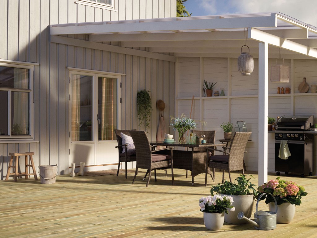 Grå treterrasse med hvit pergola, mørkt kurvbord med stoler, blomsterpotter og grill.