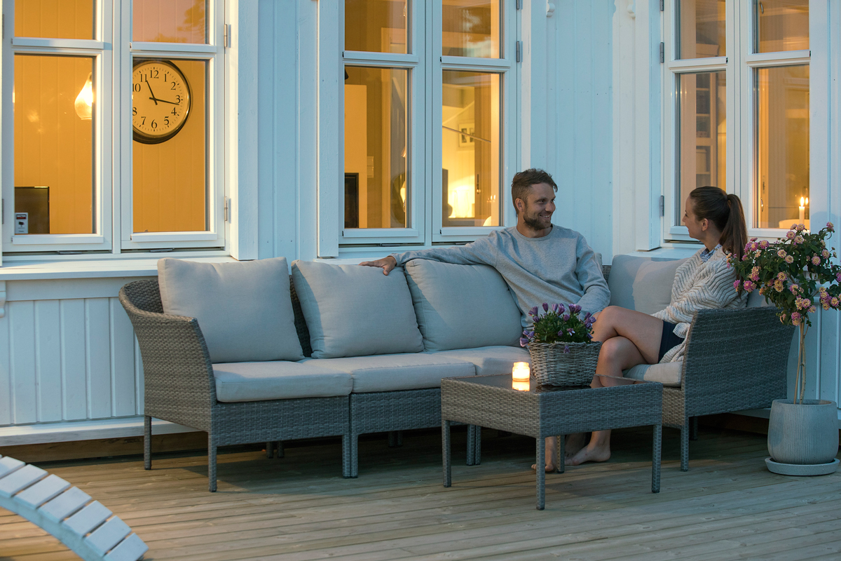 Par på grå rottingsofa på treterrasse foran hvitt hus med gylne vinduslys, stearinlys og blomster.