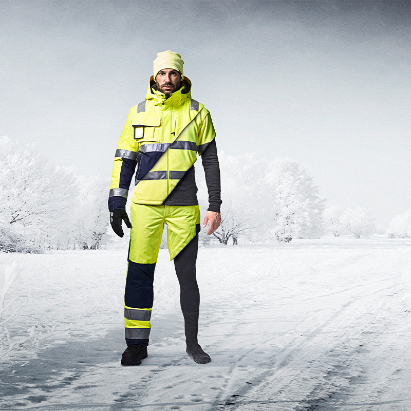 Person med gul refleksjakke og bukse og svart termo på én side, på snødekt vei