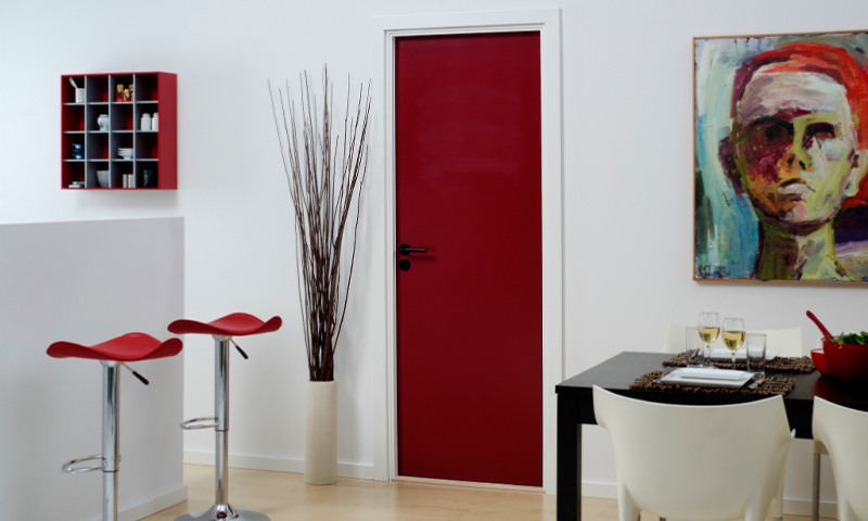 Hvitt interiør med rød dør, røde barkrakker, høy kvistvase, svart spisebord og hvite stoler.