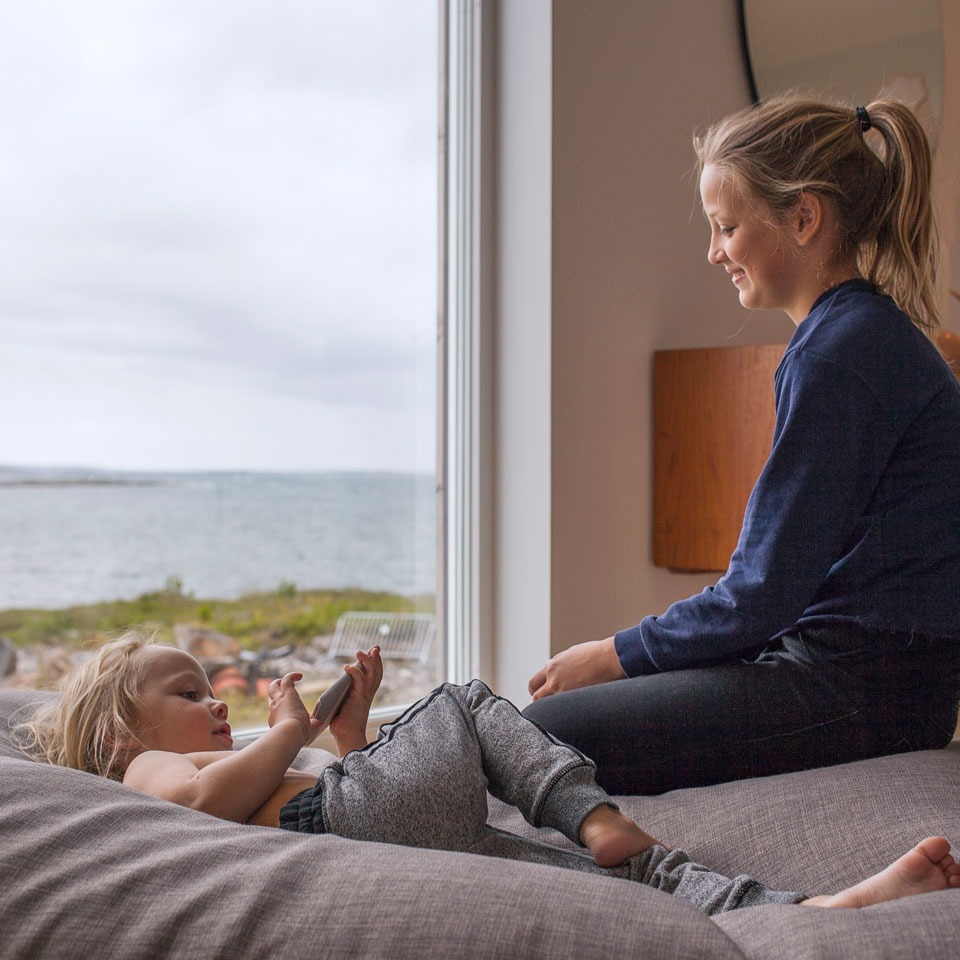 Sittende kvinne og liggende barn på grå sofa ved vindu med sjøutsikt, nøytrale fargetoner