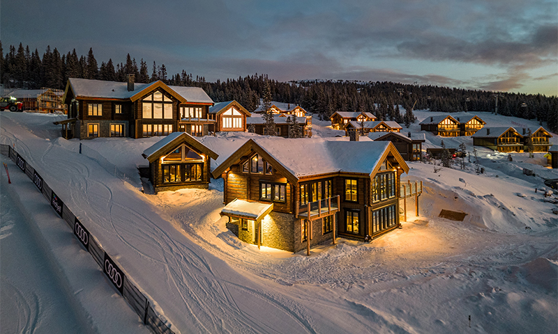 Snødekte, opplyste tømmerhytter ved skibakke og skog i fjellskumring