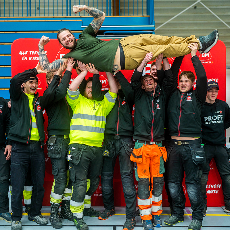 Arbeidere i gul, svart og oransje arbeidsantrekk løfter kollega i grønn t-skjorte.