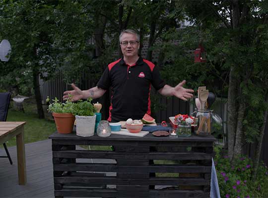 Mann i svart-rød skjorte bak svart pallebar i grønn hage med urtepotter, skåler og kjøkkenredskaper