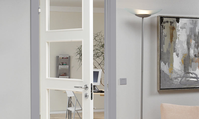 Åpen hvit dør til lyst kontor med laptop, grønn plante, stålampe og abstrakt gråmaleri.