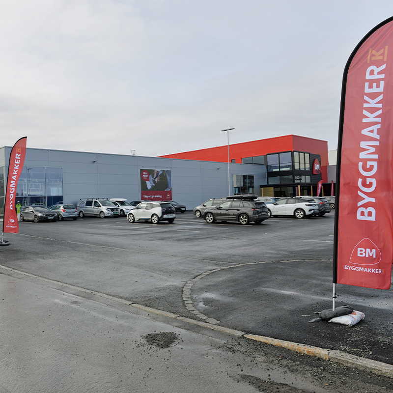 Grå Byggmakker-butikk med rødt tak, røde reklameflagg og parkerte biler foran.