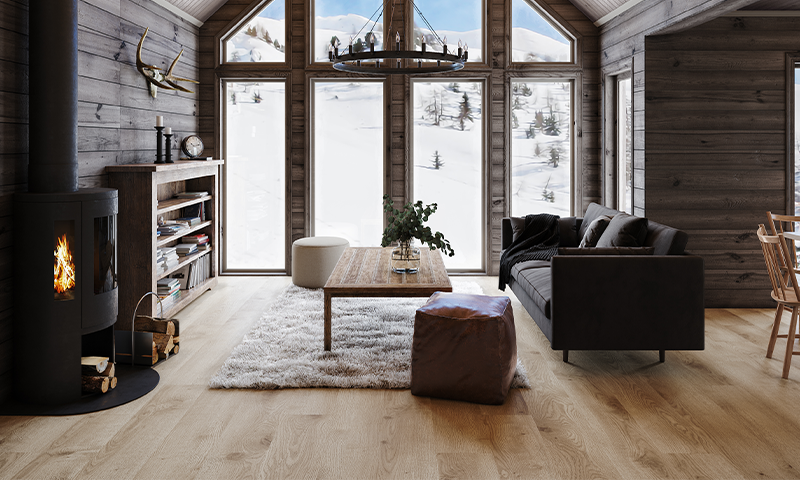 Rustikk tømmerstue med peis, mørk sofa, trebord og snødekt fjellutsikt gjennom høye vinduer