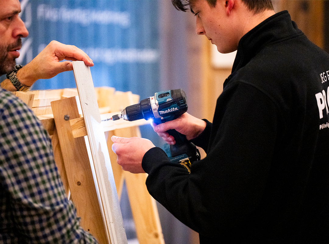 Ung person i svart genser bruker en Makita-drill på en trekonstruksjon, veiledet av en voksen mann.