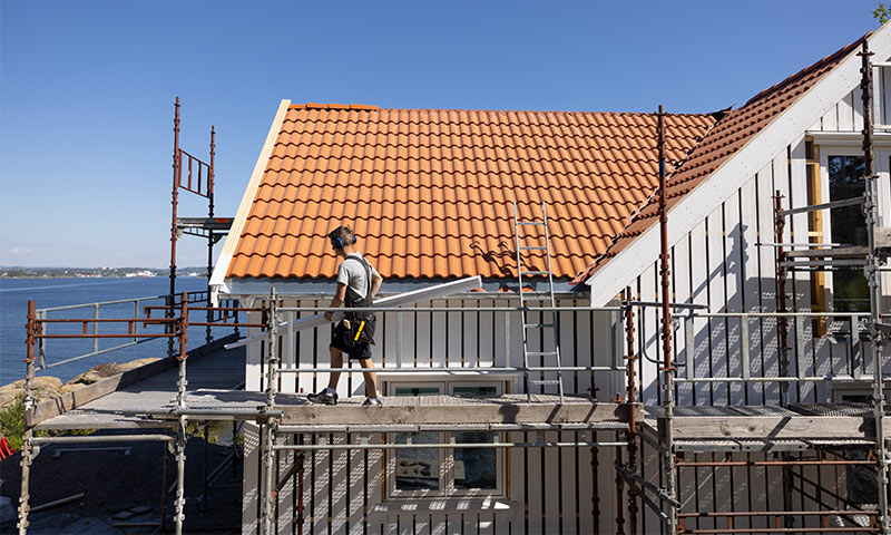 Bygningsarbeider bærer plater på stillas rundt et hvitt hus med oransje tak ved sjøen.