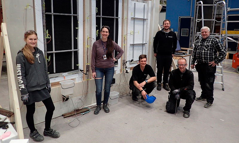 Gruppe i verksted foran hvit vegg med kabler, bøtter og verktøy, grått gulv og blå bakgrunn