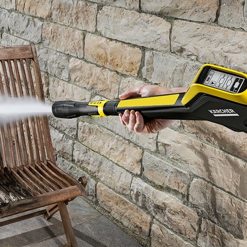 Hånd holder gul-svart Karcher høytrykksvasker som spyler trestol foran steinmur.