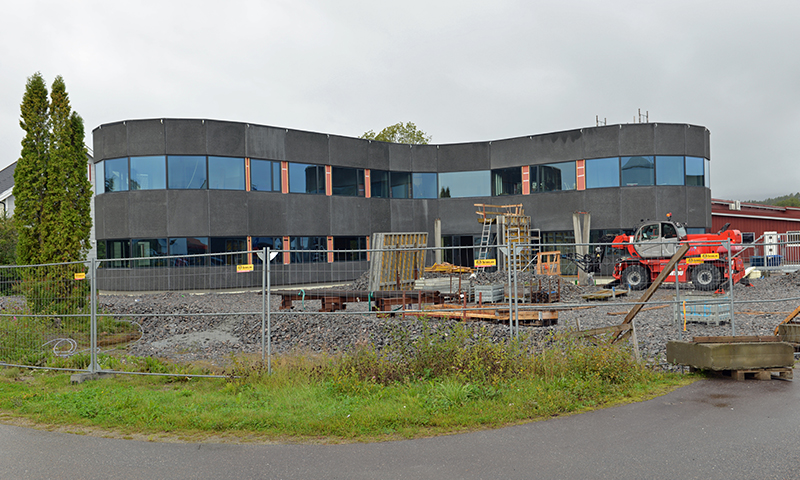 Mørk, buet kontorbygning under bygging, omkranset av gjerder, grus og rød maskin