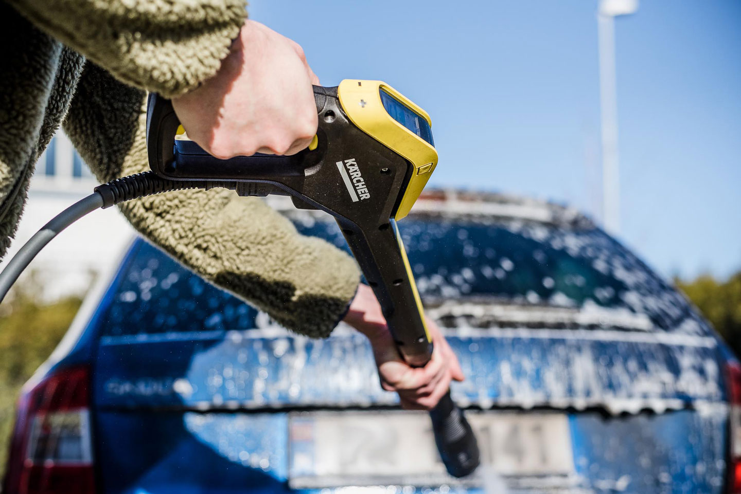 Hånd holder gul-svart Kärcher høytrykkspyler som sprayer såpevann på blå bil