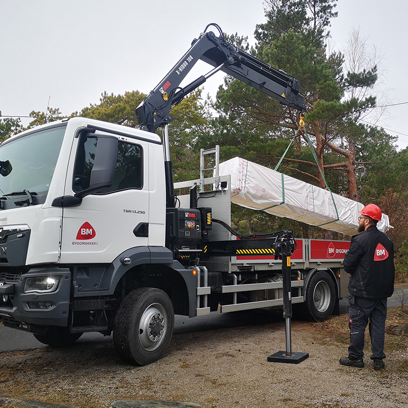 Hvit BM-lastebil med kran løfter innpakket trelast, mann i svart jakke og rød hjelm står ved siden.